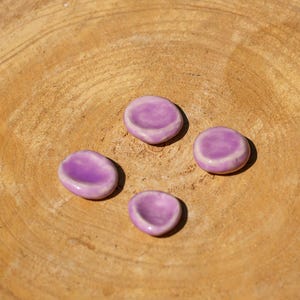 May include: Four small, lavender-colored ceramic pieces with a smooth, glossy finish. The pieces are oval-shaped with a slight indentation, resting on a light brown, wooden surface. The objects are evenly spaced, creating a simple, yet elegant composition.