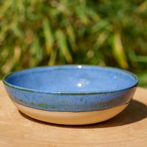 Peut inclure: Un bol en céramique fait à la main, avec un intérieur bleu clair et un extérieur de couleur crème. Le bol est rond et repose sur une surface en bois, avec un arrière-plan vert flou.