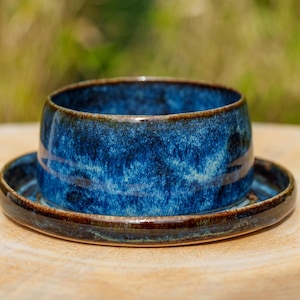 Puede incluir: Un juego de cuenco y platillo de cerámica hecho a mano. El cuenco es de color azul oscuro con un esmalte texturizado, colocado sobre un platillo a juego. El borde de ambas piezas tiene un acento marrón oscuro. El conjunto se exhibe sobre una superficie de madera clara.