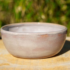 May include: A handmade ceramic bowl with a soft, muted pink and beige color palette. The bowl has a rounded shape with a slightly textured surface and a subtle, natural glaze. The rim features a darker, contrasting tone, adding visual interest.