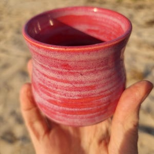 May include: A handmade, red ceramic mug with a wide, curved body and a slightly flared rim. The mug has a textured, marbled appearance with lighter pink and white streaks. The mug is held in a hand, with a blurred beach background.