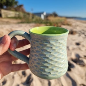 May include: A ceramic mug with a textured, scale-like pattern on the exterior in shades of blue and green. The mug features a sturdy handle and a smooth, bright lime green interior glaze, creating a distinct contrast with the matte outer finish.