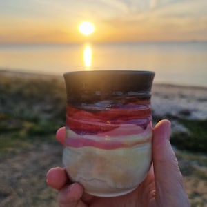 May include: A handmade ceramic cup with a layered design of cream, pink, red, and brown hues. The cup is held in a hand, with a sunset over the ocean in the background. The cup is approximately 10 cm tall.