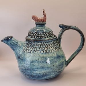May include: A handmade ceramic teapot with a blue and turquoise glaze. The lid has a unique, raised handle in a reddish-brown colour. The teapot has a textured pattern and a curved handle.