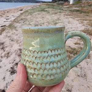 May include: A handcrafted ceramic mug with a textured design and a seafoam green glaze. The mug features a rounded handle and a light blue interior. The mug is held against a backdrop of a beach with sand and water.