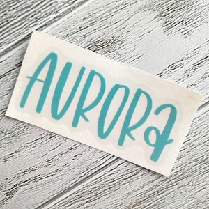 May include: A teal-colored vinyl decal with the word "AURORA" in a cursive font. The decal is on a white backing, set against a light gray, wood-grain background. The word is in all capital letters.