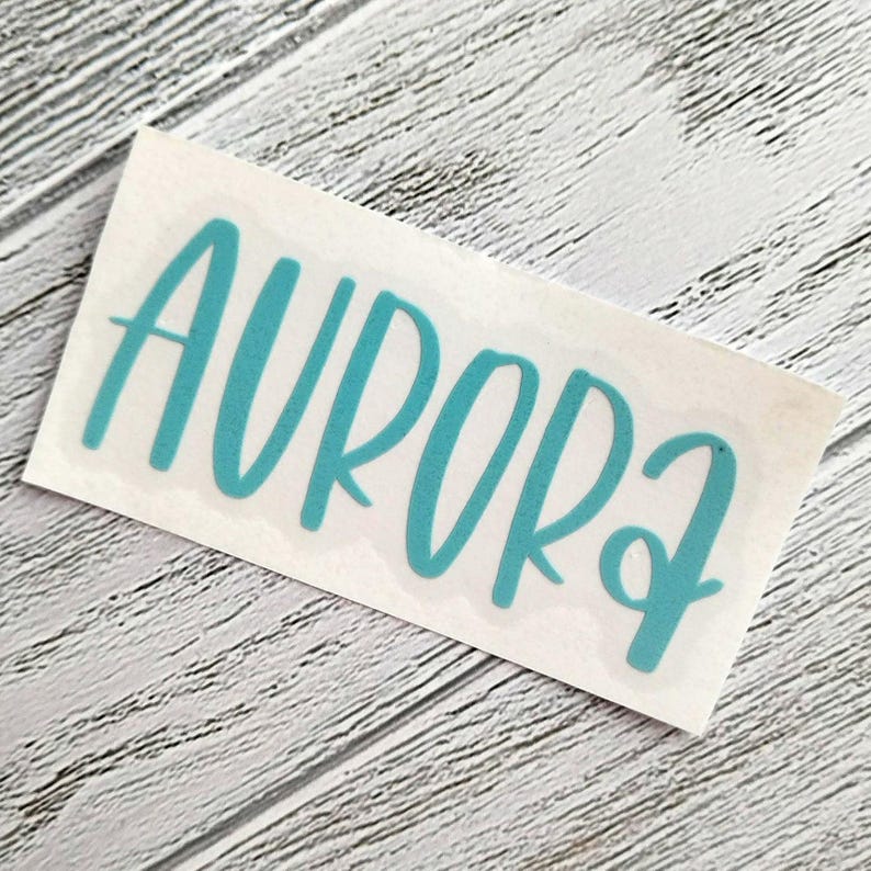 May include: A teal-colored vinyl decal with the word "AURORA" in a cursive font. The decal is on a white backing, set against a light gray, wood-grain background. The word is in all capital letters.