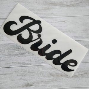 May include: A white sticker with the word "Bride" in a bold, black, cursive font. The sticker is rectangular and is placed on a light gray wooden surface.