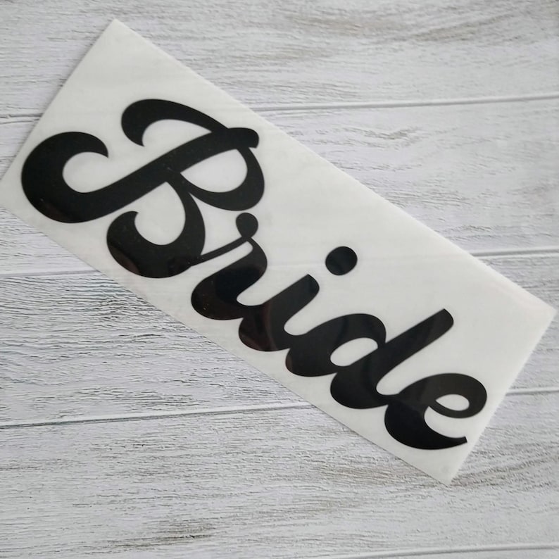 May include: A white sticker with the word "Bride" in a bold, black, cursive font. The sticker is rectangular and is placed on a light gray wooden surface.