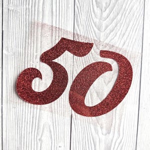 May include: A glittery red number 50 cutout featuring a bold, stylised font with a textured, sparkling surface. The number is crafted from a single piece of material, showcasing a deep crimson hue that catches the light across its entire surface.