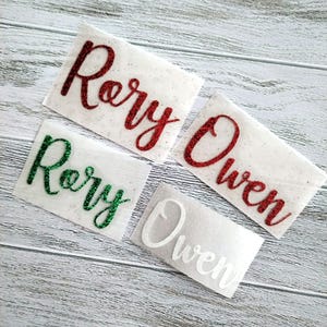 May include: Four personalised name decals with glitter. The names are Rory and Owen. Two of the names are in red glitter, one is in green glitter, and one is in white.