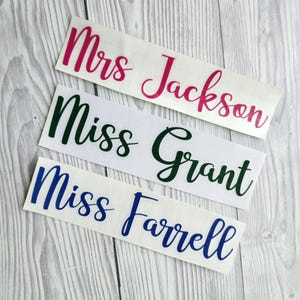 May include: Three name decals with cursive lettering. The top decal reads "Mrs Jackson" in pink. The middle decal reads "Miss Grant" in dark green. The bottom decal reads "Miss Farrell" in blue. All decals are on a white background.