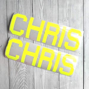May include: Two neon yellow vinyl decals, each displaying the name "CHRIS" in a bold, sans-serif font. The rectangular decals are on a light grey backing, set against a backdrop of light grey wooden planks.