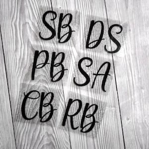 May include: Black cursive letters SB, DS, PB, SA, CB, and RB are arranged on a light wood grain background. The letters are on a transparent backing. The letters are likely for a craft project or personalised item.