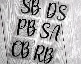 May include: Black cursive letters SB, DS, PB, SA, CB, and RB are arranged on a light wood grain background. The letters are on a transparent backing. The letters are likely for a craft project or personalized item.