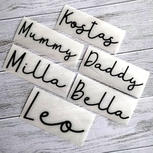 May include: White rectangular labels with black cursive text. The labels include the names "Mummy", "Milla", "Leo", "Kostas", "Daddy", and "Bella". The labels are arranged on a light wood-grain surface.