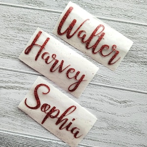 May include: Three white labels with red glitter lettering. The labels read "Harvey", "Walter", and "Sophia".