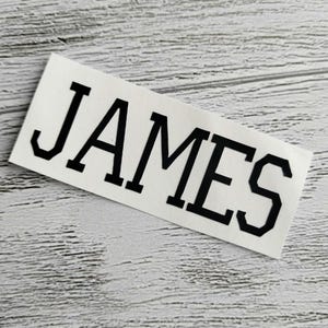 May include: A white sticker with the name "JAMES" printed in black block letters.