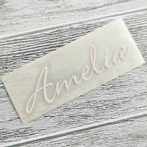 May include: A light pink decal with the name "Amelia" in a cursive font. The decal is on a clear backing, set against a light wood-grain surface. The script is elegant and flowing, suitable for personalization.