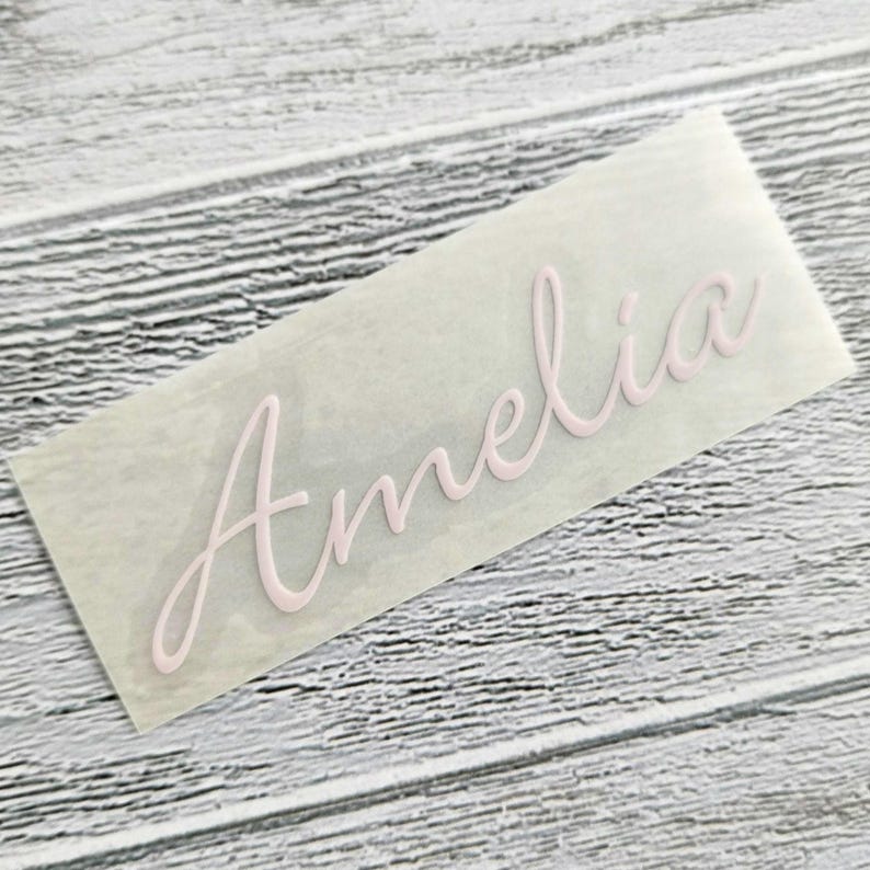 May include: A light pink decal with the name "Amelia" in a cursive font. The decal is on a clear backing, set against a light wood-grain surface. The script is elegant and flowing, suitable for personalization.