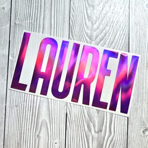 May include: A rectangular adhesive decal featuring the name LAUREN in bold, capital, sans-serif letters. The lettering displays a vibrant, iridescent finish with shifting gradients of deep purple, magenta, and bright pink hues across the surface.