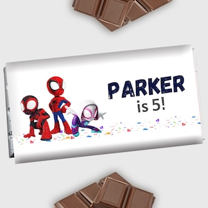 May include: A white chocolate bar wrapper with a Spiderman theme. The wrapper features three Spiderman characters, including Spiderman, Miles Morales, and Gwen Stacy. The text on the wrapper reads "PARKER is 5!"