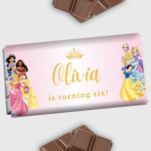 May include: Pink and gold princess-themed chocolate bar wrapper with a gold crown and the text "Olivia is turning six!"  The wrapper features a group of Disney princesses, including Snow White, Cinderella, and Belle.