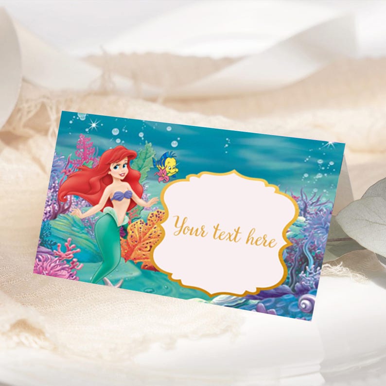 May include: A place card featuring Ariel, the Little Mermaid, with a blank space for writing a name. The card has a blue and green background with coral and seaweed.