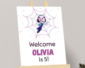 May include: A white sign on a wooden easel features a cartoon character in a spider web design. The sign reads "Welcome OLIVIA is 5!" in purple and brown text. The background is white with small colorful dots.