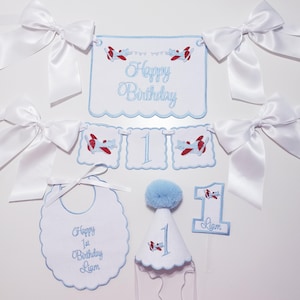 Może przedstawiać: Zestaw białych, haftowanych dekoracji urodzinowych z niebiesko-czerwonymi wzorami samolotów. Zawiera baner z napisem "Happy Birthday", baner z cyfrą "1", śliniaczek, czapeczkę urodzinową i topper na tort. W zestawie również białe kokardy.