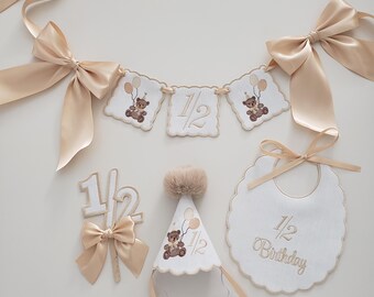 May include: A collection of baby's first birthday decorations in cream and beige. Includes a banner with teddy bear and balloon embroidery, a bib, a party hat, and a cake topper. The banner and bib read "1/2 Birthday".