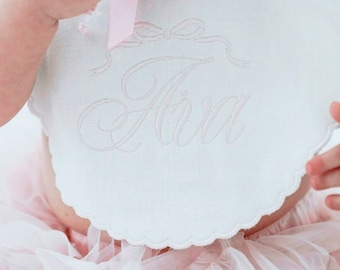 May include: White bib with a scalloped edge and pink satin ribbon bow. The bib is embroidered with the name "Ava" in pink thread.