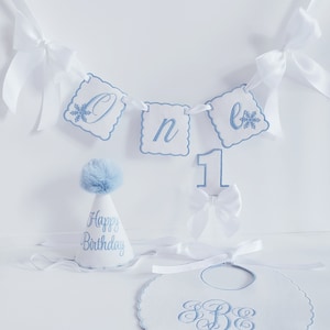 May include: A white and blue baby's first birthday party set with a banner that says "One", a party hat that says "Happy Birthday", and a bib with the monogram "S.B.E."