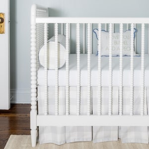 Puede incluir: Una cuna blanca con un diseño de tablero de cuentas, con una sábana bajera blanca y una falda de cuna a cuadros grises y blancos. Se puede ver una almohada decorativa con el texto "I LOVE YOU A BUSHEL AND A PECK". También hay un cojín cilíndrico redondo.