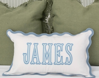 Pode incluir: Travesseiro branco com uma borda azul festoneada e o nome "JAMES" bordado em azul na frente.