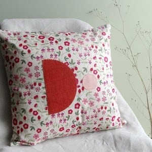 Könnte beinhalten: Ein weißes Kissen mit einem Blumenmuster mit rosa und roten Blüten. Das Kissen hat einen großen roten Halbkreis und einen kleineren rosa Kreis, die auf die Vorderseite genäht sind.