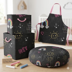 Könnte beinhalten: Ein Set von Spielzimmer-Accessoires für Kinder, darunter zwei schwarze Aufbewahrungswürfel mit rosa Seilgriffen, ein runder Pouf und eine Schürze. Jeder Artikel zeigt farbenfrohe Kreidezeichnungen von Regenbogen, Sonnen und Hüpfkästchen. Das Wort "HEY" steht auf einem Würfel.