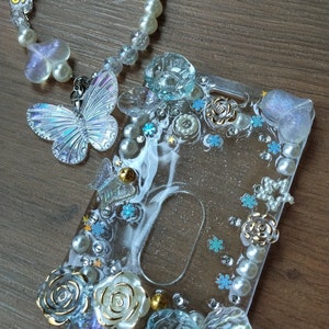 May include: A clear plastic ID badge holder with a white background and a variety of decorative elements, including blue and white flowers, pearls, and butterflies. The holder is decorated with glitter and has a clear window for displaying an ID card.