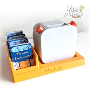 May include: A white projector with a red button on top, sitting on a yellow stand with the word "STORIES" printed on it. The stand has a slot for cards, which are visible in the image. The cards have colourful illustrations and text, including "OUR PLANET" and "Tropical Rainforests".
