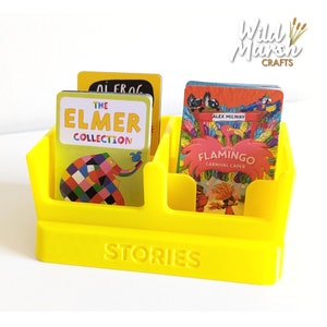 May include: Yellow plastic story card holder with the words "STORIES" on the front. The holder has two compartments and is filled with colourful story cards featuring the characters "Elmer" and "Hotel Flamingo".