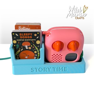 May include: A light blue plastic organiser with the text STORY TIME embossed on the front. It holds a stack of small rectangular cards, including one labelled SLEEPY SONGS CASPAR BABYPANTS, and a pink portable audio player with two orange knobs.