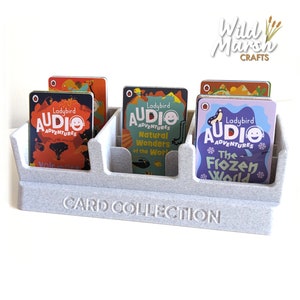 May include: A grey card holder with the text "CARD COLLECTION" printed on the front. The holder has three slots and is filled with Ladybird Audio Adventures cards. The cards feature colourful illustrations and the text "Ladybird Audio Adventures".