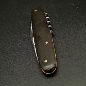 Op de afbeelding: Een inklapbaar zakmes met een donkerbruin handvat en twee koperen pinnen. Het handvat heeft een gebogen vorm met een metalen rand en een zichtbaar kurkentrekker-mechanisme langs de zijkant van het frame, wat een compact ontwerp laat zien.