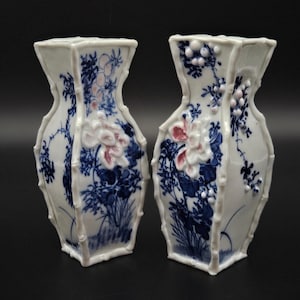 May include: A pair of white porcelain vases with a square, bamboo-like frame. Each vase features blue painted floral patterns and raised, three-dimensional white and pink flower accents. The design includes intricate blue botanical illustrations.
