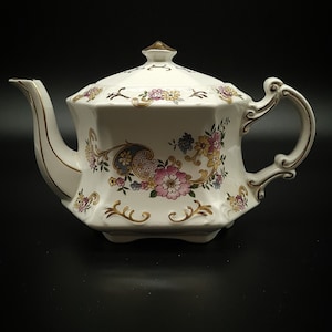 May include: A white teapot with gold trim and a floral pattern. The teapot has a lid with a gold knob and a spout on the side. The teapot is decorated with pink, blue, and yellow flowers.