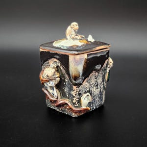 Może przedstawiać: Kwadratowy ceramiczny słoik z pokrywką i ciemnym, nakrapianym szkliwem. Na pokrywce znajduje się mała, wyrzeźbiona figurka małpy. Boki słoika ozdobione są reliefami małp oraz teksturowanymi, ziemistymi wzorami w odcieniach brązu, czerni i kremu.