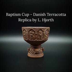 Op de afbeelding: Een bruine terracotta doopbeker replica van L. Hjorth. De beker heeft gedetailleerde reliëfs van figuren en scènes rond de kom en de basis. De tekst "Baptism Cup - Danish Terracotta Replica by L. Hjorth" is weergegeven.