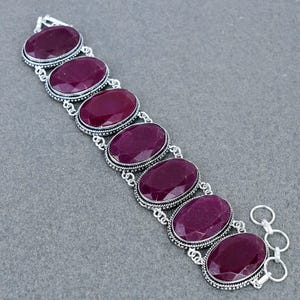 Puede incluir: Una pulsera con siete piedras ovaladas de color magenta intenso engastadas en marcos de metal plateado. Cada piedra está rodeada por un borde trenzado. Los eslabones están unidos por pequeños anillos plateados y terminan en un cierre.