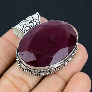 Pode incluir: Um pingente de prata com uma gema de cor vermelha escura de forma oval e facetada, engastada em uma moldura de prata. O pingente tem uma fivela de prata adornada.
