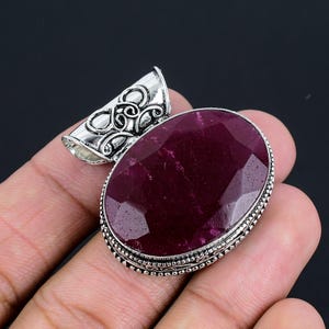 Puede incluir: Un colgante de plata con una piedra preciosa ovalada facetada de color morado oscuro. El colgante tiene una baíl de plata adornada con un diseño decorativo.